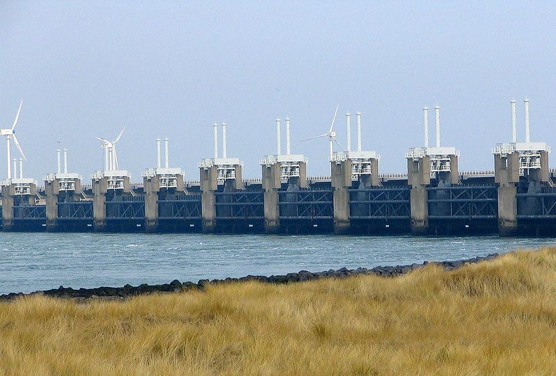 The Eastern Scheldt Storm Surge barrier sits in the sea, beyond a yellow grass covered shore.