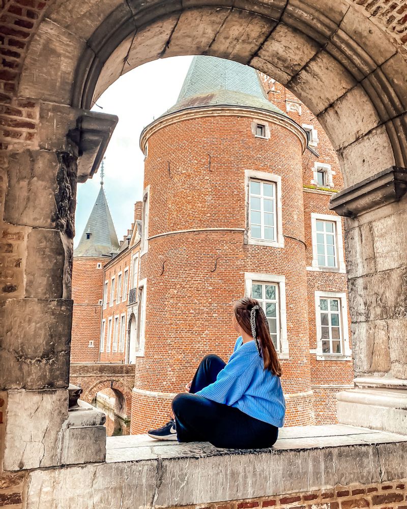 A woman in a blue jumper sits in a large brick archway overlooking Alden Biesen Castle in Bilzen, Belgium.