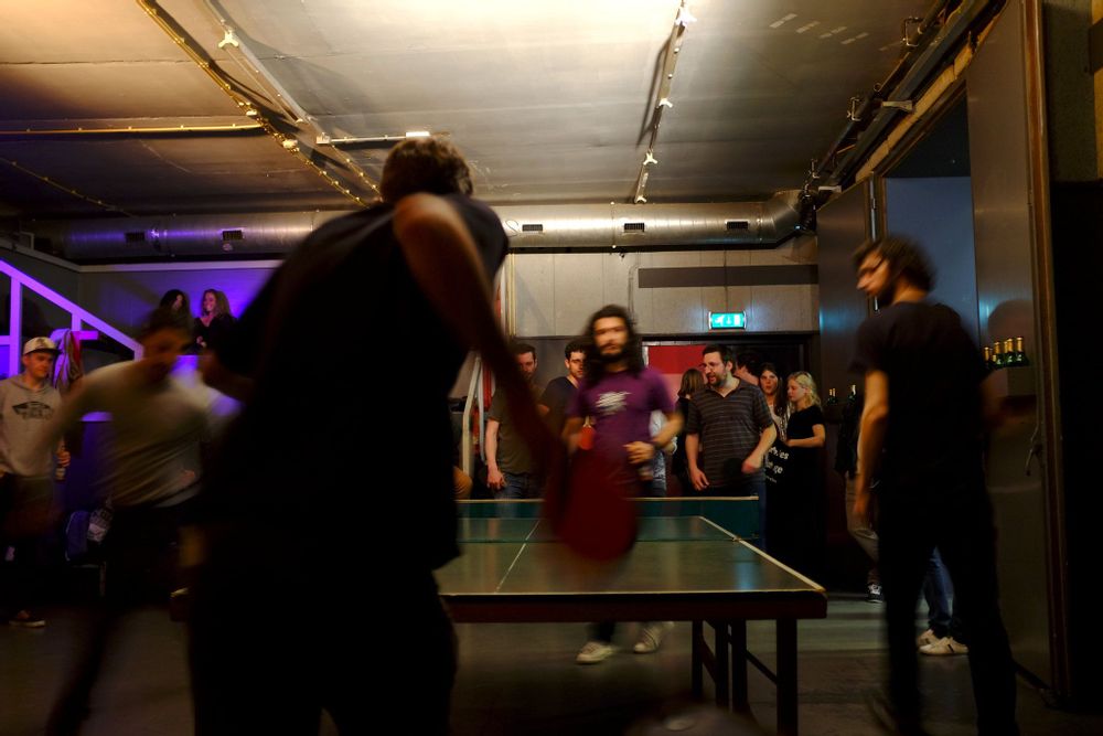 A room crowded with people onlooking two men playing ping pong at OT301