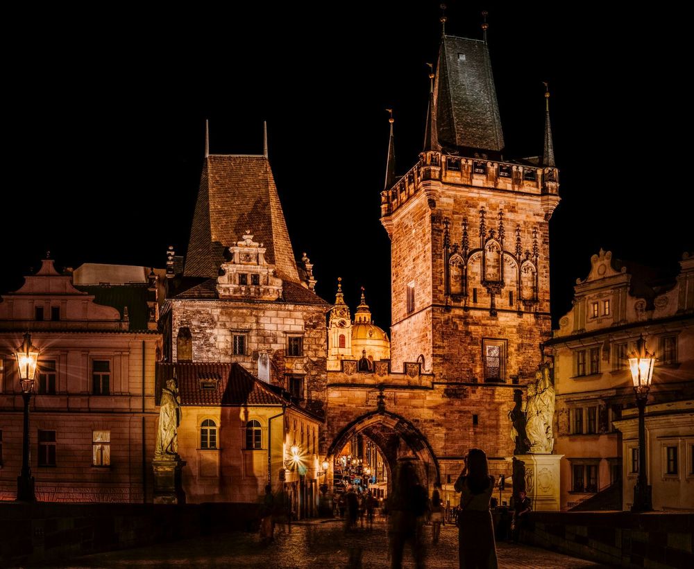 Women taking photos of an arch and surrounding buildings at night by Prague Castle, Czechia