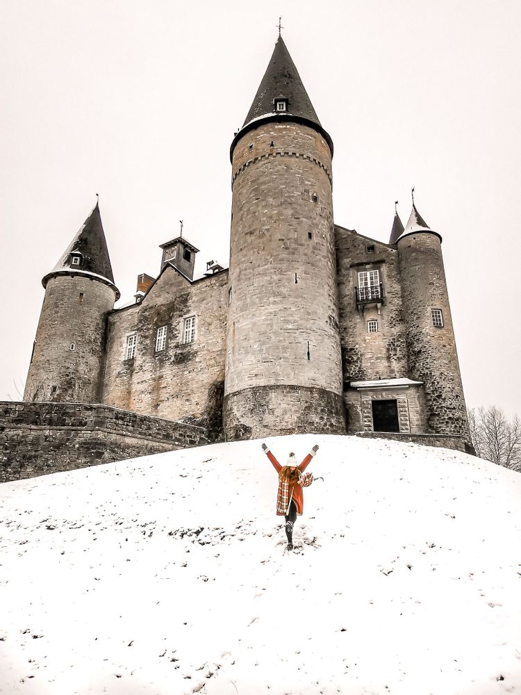 Woman wearing an orange coat and scarf standing in the snow in front of Veves Castle, Belgium