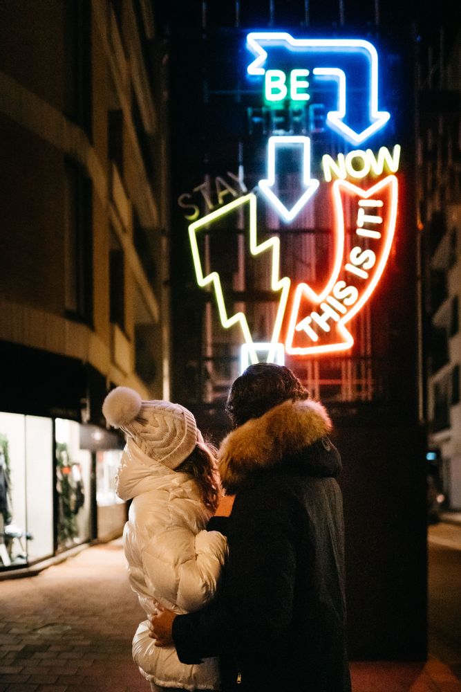 Father and toddler viewing neon light art display that reads “Be Now This Is It” at the Light ART festival, Knokke-Heist