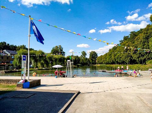 A view of Lake Conchibois with people swimming and sitting on the edge. Two lifeguards are sitting under an umbrella.