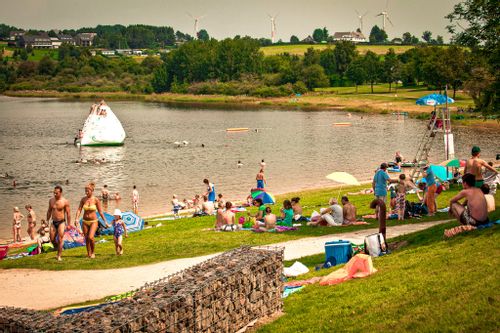 People sitting on the banks of Lake Butgenbach at VENNtastic beach, with a lifeguard in a tower and inflatables in the water.