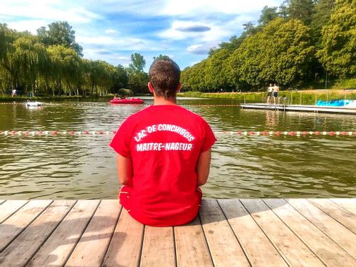 A man wearing a lifeguard shirt sitting on the edge of the swimming area at Lake Conchibois, Saint-Léger.